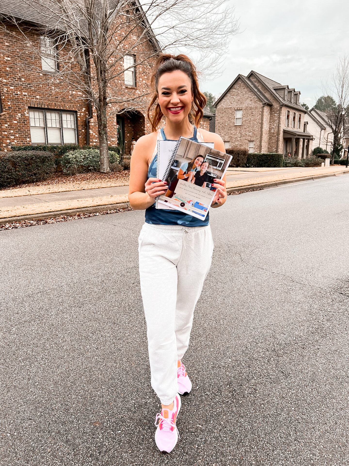 5 Tips To Staying On Track With Health Goals During Busy Seasons - Heather is wearing athleisure and holding her meal planning books 