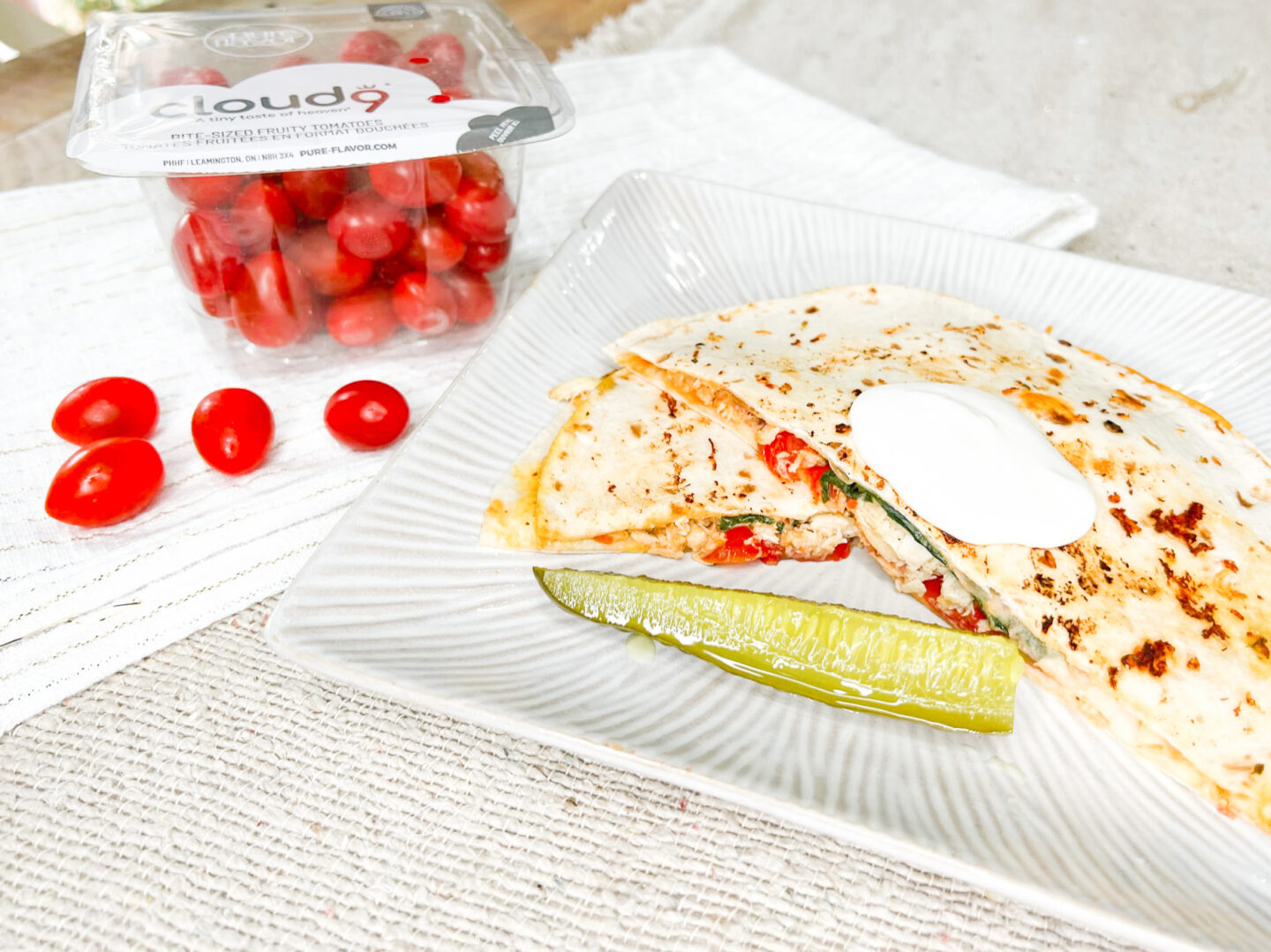A low-carb tomato basil chicken wrap on a white plate with a pickle. A container of Cloud 9 grape tomatoes is in the background. 